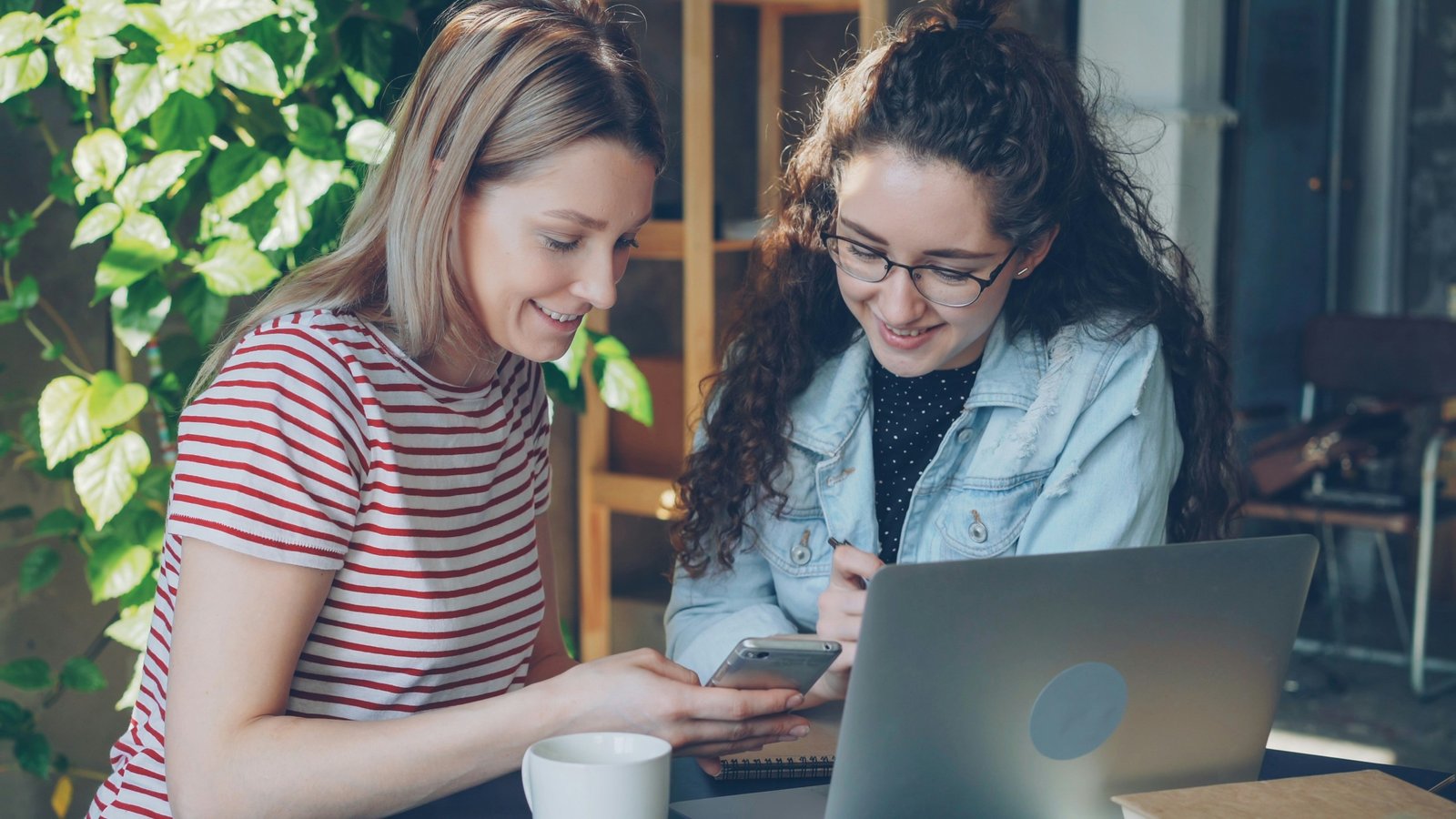 Close-up of two female students using smart phone while sitting together. Attractive blond girl pointing touchscreen and chatting with her friend. Modern technology for young people concept.